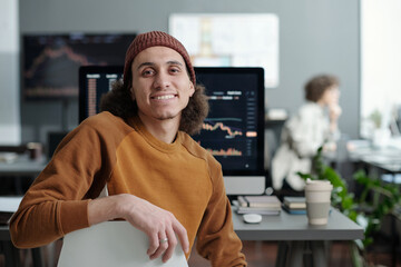 Poster - Young smiling IT support manager sitting by workplace in front of camera against computer monitor with graphic data on screen