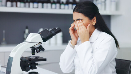 Wall Mural - Female scientist looking through microscope and rubbing her eyes while suffering from eye strain or discomfort. Stressed biotechnology specialist does analysis of test sample in medical lab