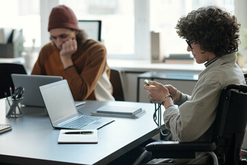 Aufkleber - Side view of young businesswoman with disability scrolling in mobile phone while sitting in wheelchair in front of laptop and male colleague