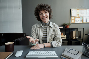 Poster - Young smiling female manager in casualwear sitting by desk in front of computer moniot, looking at screen and communicating in video chat