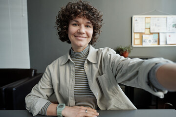 Poster - Smiling brunette businesswoman with dark short curly hair looking at camera while holding mobile gadget in front of herself