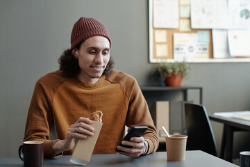 Aufkleber - Young smiling multi-ethnic male employee with smartphone having sandwich and communicating in video chat at lunch break