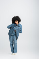 Wall Mural - full length of curly african american woman in denim outfit looking at camera while standing on grey.