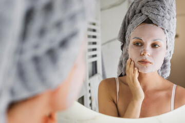 Wall Mural - Woman in bathroom with applied sheet mask on her face looking in the mirror