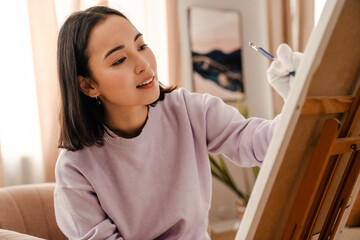 Wall Mural - Young asian woman artist drawing with pencil on canvas while sitting at home