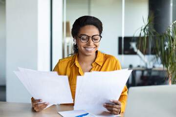 Wall Mural - Excited black businesswoman reading papers and positive business report, sitting at workplace in modern office interior