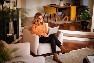 Poster - Mid aged woman using earphones and notebook and sitting in an armchair at home