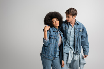 young man in denim outfit hugging curly african american woman isolated on grey.