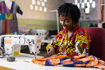 African woman fashion tailor wearing ankara dress with locs hair sewing an outifit in her dressmaking studio with an electric machine.