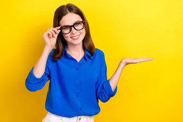 Poster - Photo of young satisfied successful lady wear blue shirt new eyeglasses hold palm promotion new business center isolated on yellow color background