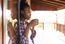 African American Woman Spending Time In Log Cabin Drinking Coffee On Balcony