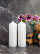 Two white candles on a dark marble table with a bouquet of flowers and a teddy bear.