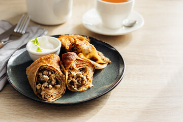 Wall Mural - Crepes stuffed with chicken and mushrooms with sour cream and cup of tea on the wooden table.