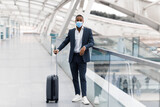 Fototapeta  - Black Man Wearing Suit And Medical Mask Posing With Suitcase At Airport