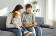 Parents and their adorable toddler baby reading book. Happy family sitting on couch in cozy living room having good time together. Babies early development, indoor activities