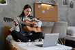 Attractive asian teenager girl learning to play guitar using laptop computer sitting on bed at home.