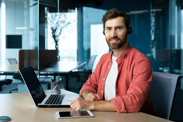 Wall Mural - Portrait of mature businessman in red shirt inside office, man with beard and video call headset smiling and looking at camera, online customer support worker with laptop.