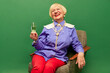Happy old woman, grandmother in stylish colorful clothes sitting with legs into bowl and drinking champagne on green studio background. Concept of age, fashion, lifestyle, emotions, facial expression