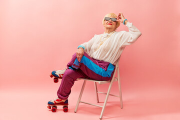 Relaxed vibe. Beautiful old woman, grandmother in stylish sportive trousers posing on vintage rollers over pink studio background. Concept of age, fashion, lifestyle, emotions, facial expression