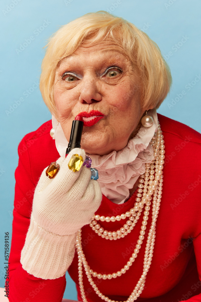 Applying lipstick. Beautiful old woman, grandmother in stylish red dress  and pearl necklace posing over blue studio background. Concept of age,  fashion, lifestyle, emotions, facial expression Stock-foto | Adobe Stock