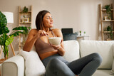 Fototapeta Panele - Beautiful young woman is sitting on the sofa in the living room and eating oatmeal