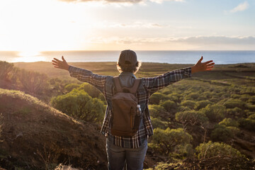 Sticker - Back view of senior caucasian woman with hat and casual clothing standing with open arms looking at the horizon over sea. Elderly lady enjoying nature and freedom admiring landscape at sunset light