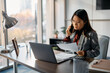 © Drazen - Asian businesswoman talking on cell phone while analyzing documents and working on laptop in office.