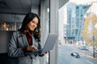 © Drazen - Happy Asian businesswoman working on laptop by window.