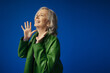 © LIGHTFIELD STUDIOS - excited grey haired woman in green leather jacket showing wow gesture and looking away isolated on blue