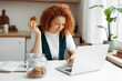 © shurkin_son - Indoor image of redhead curly young woman freelancer working on laptop sitting at kitchen table with cookie in hand and jar of cookies next to her, having coffee break. Unconscious eating