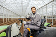 An agronomy worker is driving tractor in hothouse and giving thumbs up while smiling at the camera.