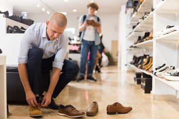 Interested man visiting shoe store in search of new shoes, choosing and trying on while sitting on pouf.