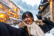 Asian woman tourist using mobile phone taking selfie during travel onsen area Ginzan Onsen in Yamagata prefecture, Japan in snow day. Attractive girl travel local village landmark on winter vacation