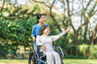 Young asian care helper with asia elderly woman on wheelchair relax together park outdoors to help and encourage and rest your mind with green nature. Hand pointing forward