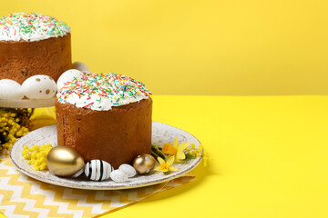 Traditional Easter cakes with sprinkles, painted eggs and beautiful spring flowers on yellow background, space for text