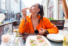 Young Woman Eating Pizza In A Street City Cafe. A Beautiful Brunette In A Bright Orange Jacket Takes A Delicious Bite.
