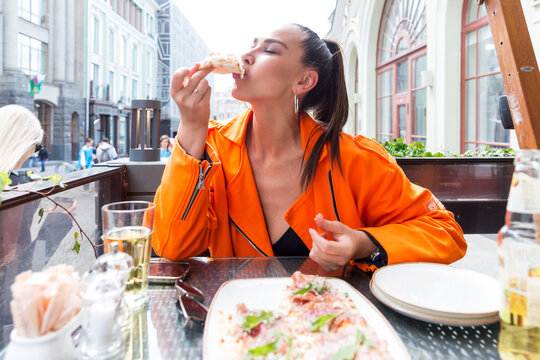 young woman eating pizza in a street city cafe. a beautiful brunette in a bright orange jacket takes