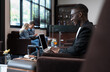 © boonsom - Man with suitcase sitting in airport waiting area. African businessman using laptop and waiting for his flight