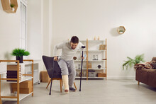Young Man With A Broken Leg In A Plaster Cast Tries To Stand Up Off His Chair And Walk With A Stick And Crutches In The Living Room At Home. Injury, Accident, Fracture Concept