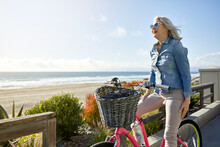 Smiling Senior Woman With Bicycle Standing At Manhattan Beach Against Sky During Sunny Day