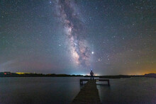 Man Standing On Dock Looking Out Over Harbor At Milky Way