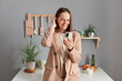 © sementsova321 - Indoor shot of smiling delighted brown haired woman wearing beige suit standing near table on kitchen at home, using cell phone, checking social networks, browsing internet.