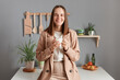 © sementsova321 - Portrait of cheerful smiling delighted young adult woman wearing beige suit standing near table on kitchen at home, drinking coffee in the morning, looking at camera.