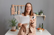 © sementsova321 - Photo of laughing positive young adult woman wearing beige suit, holding paper documents and mobile phone, looking at camera, standing near table on kitchen at home.