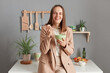 © sementsova321 - Indoor shot of smiling laughing young adult woman wearing beige suit standing near table on kitchen at home, looking at camera and laughing, eating tasty dish.