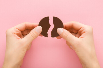 Young adult woman hands holding broken pieces from circle shape of black dark chocolate candy on light pink table background. Pastel color. Sweet snack. Closeup. Point of view shot. Top down view.