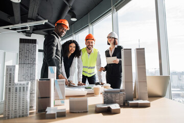 Wall Mural - Multiracial coworkers builders and architects standing near table with blueprints, gadgets and design of buildings residential project maquette and looking at camera.