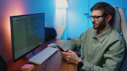 programmer man playing mobile video game by smartphone, while writing code automatically on computer