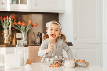 happy excited kid girl baking cake for Easter emotional child cook dessert present for mother day in white sunny kitchen Home bakery. Bottle of milk, eggs, flour are on the table