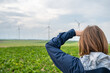 Woman with brown hair and blue jacket is looking at a wind park from the distance, wind park with wind turbines in agricultural field area, rear view